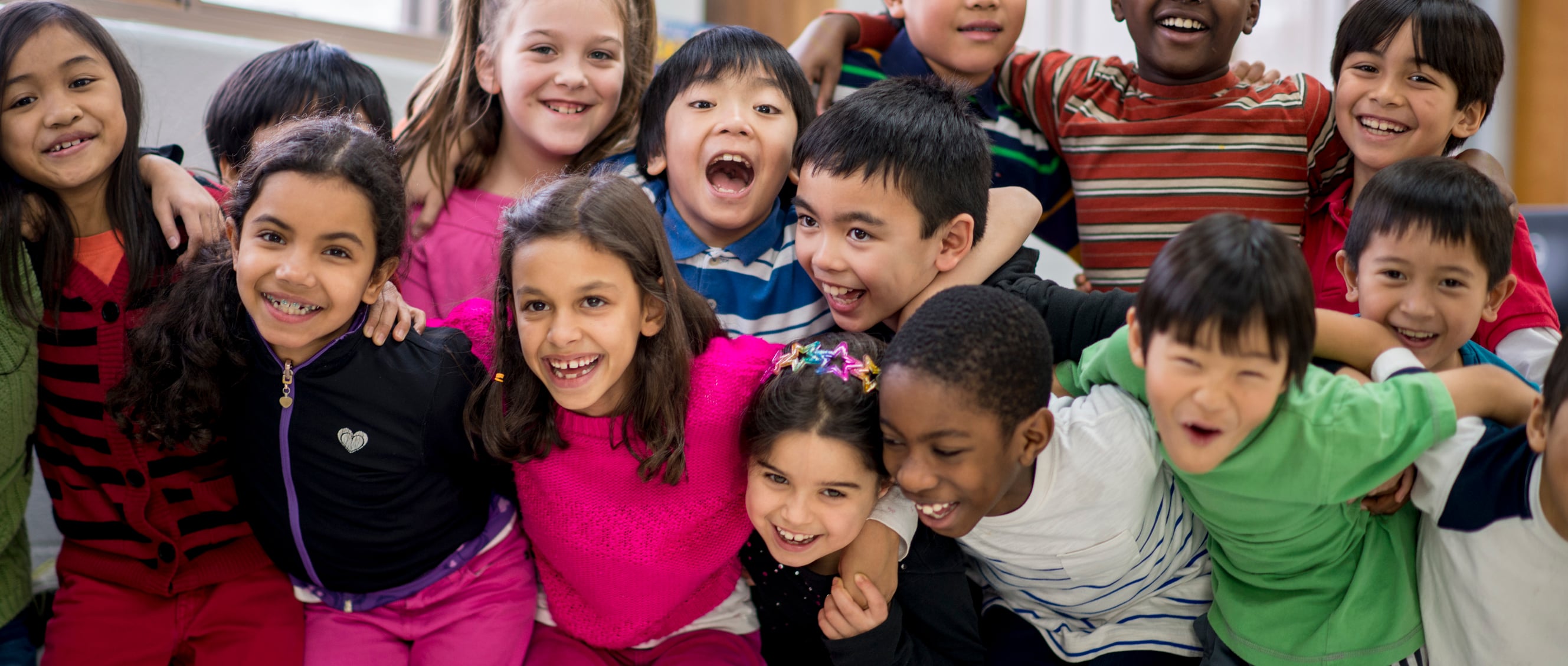 A group of smiling elementary school children of diverse backgrounds huddle together with arms around each other in a classroom.